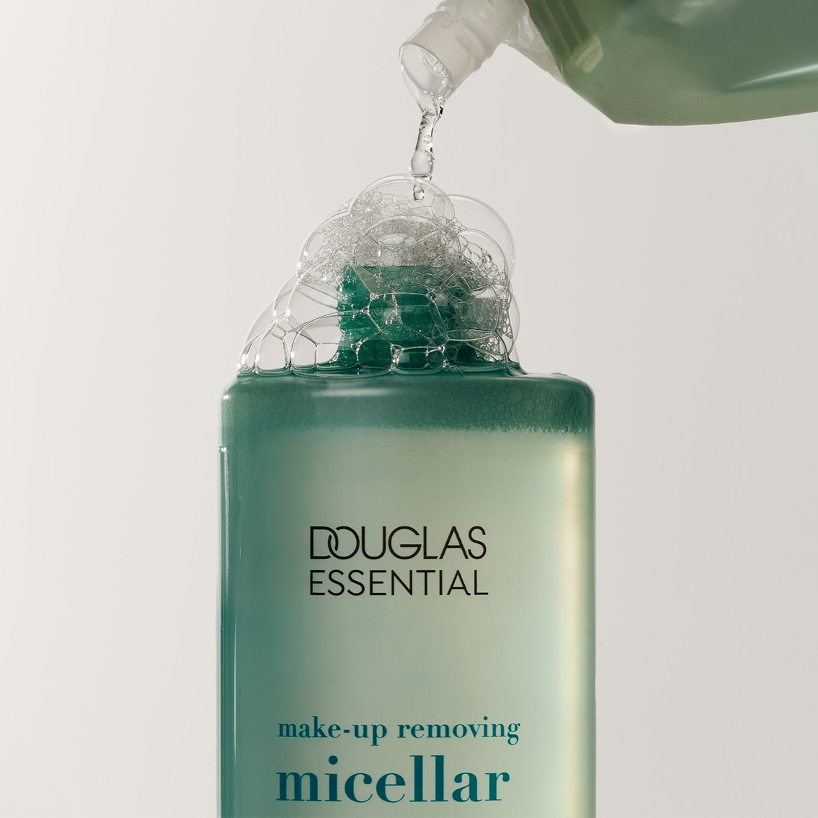Švelnus „Douglas Essential“ makiažo valiklis micelinis vanduo, supilstytas į žalsvą buteliuką, sukuria putų burbuliukus.