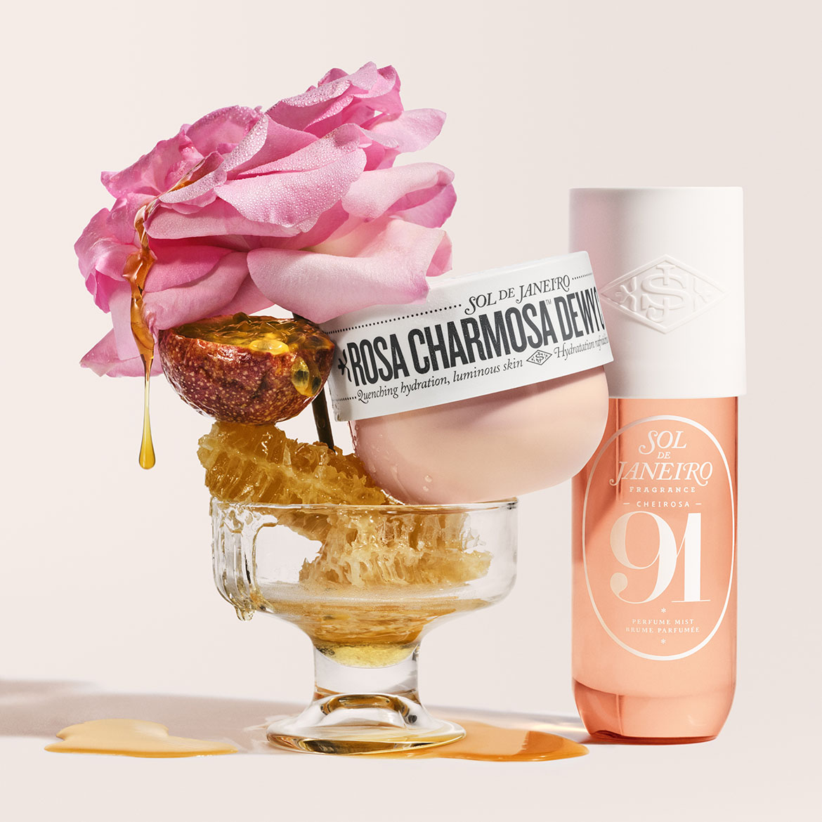 Šalia „Sol de Janeiro“ „Rosa Charmosa Dewy“ kremo, kuris drėkina ir suteikia odai švytėjimo, ir „Cheirosa '91“ purškiklio, stiklinėje taurėje pavaizduotas rožinis žiedas, pasifloros vaisius ir korėtas medus.