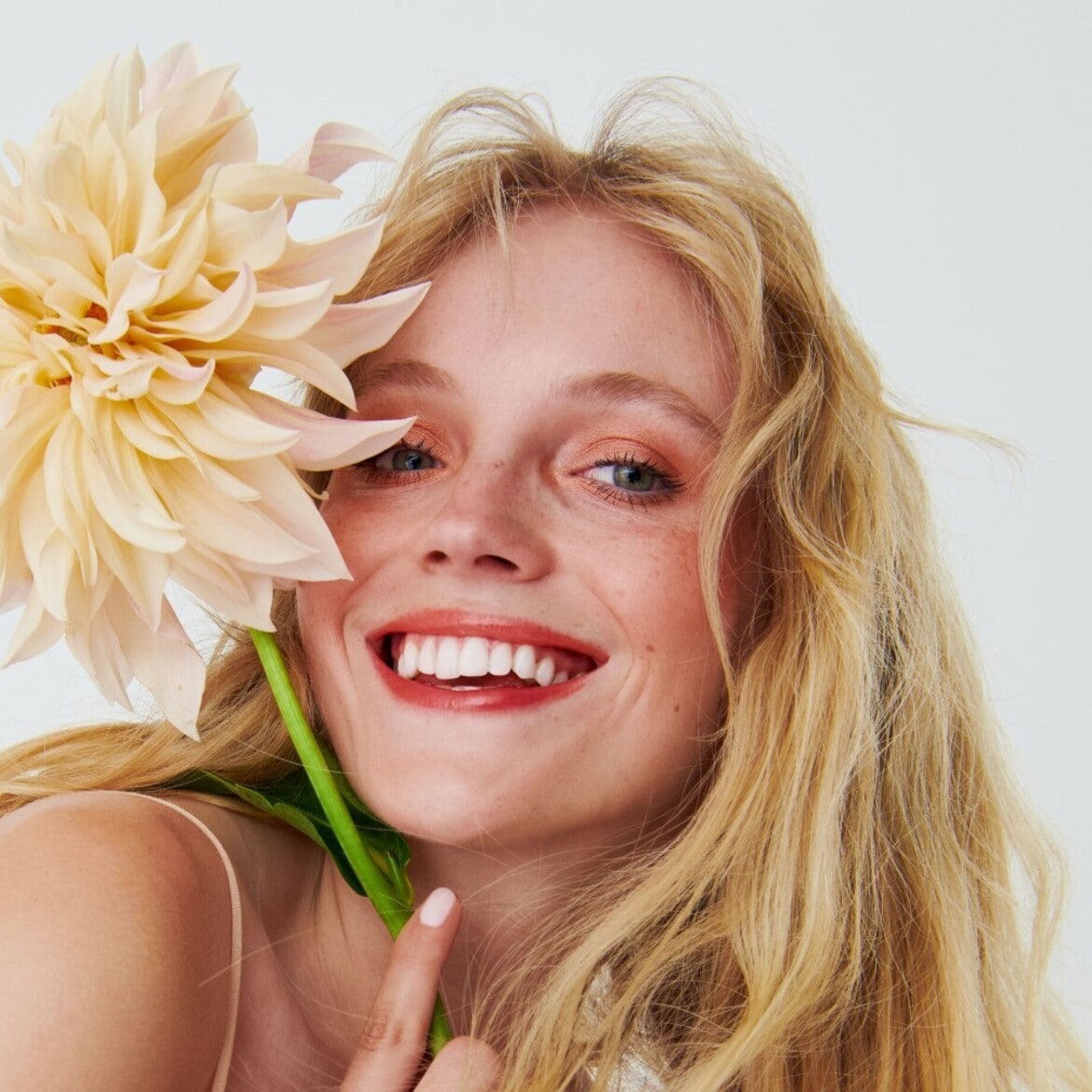 Švytinti šypsena ir šviesūs plaukai, papuošti kreminės spalvos žiedu, sukuria gaivumo ir natūralumo įspūdį.