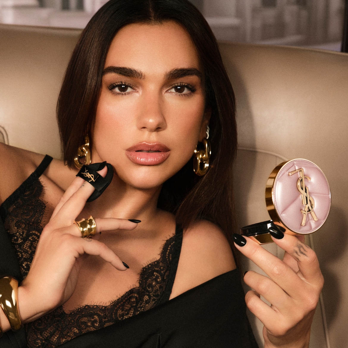 Dua Lipa sėdi ant smėlio spalvos kėdės, vilkėdama juodą nėriniuotą palaidinę, su juodais nagais, laiko atidarytą šviesiai rožinės spalvos kompaktinę pudrą su auksiniu YSL logotipu ir juodą kempinėlę su auksiniu YSL logotipu.