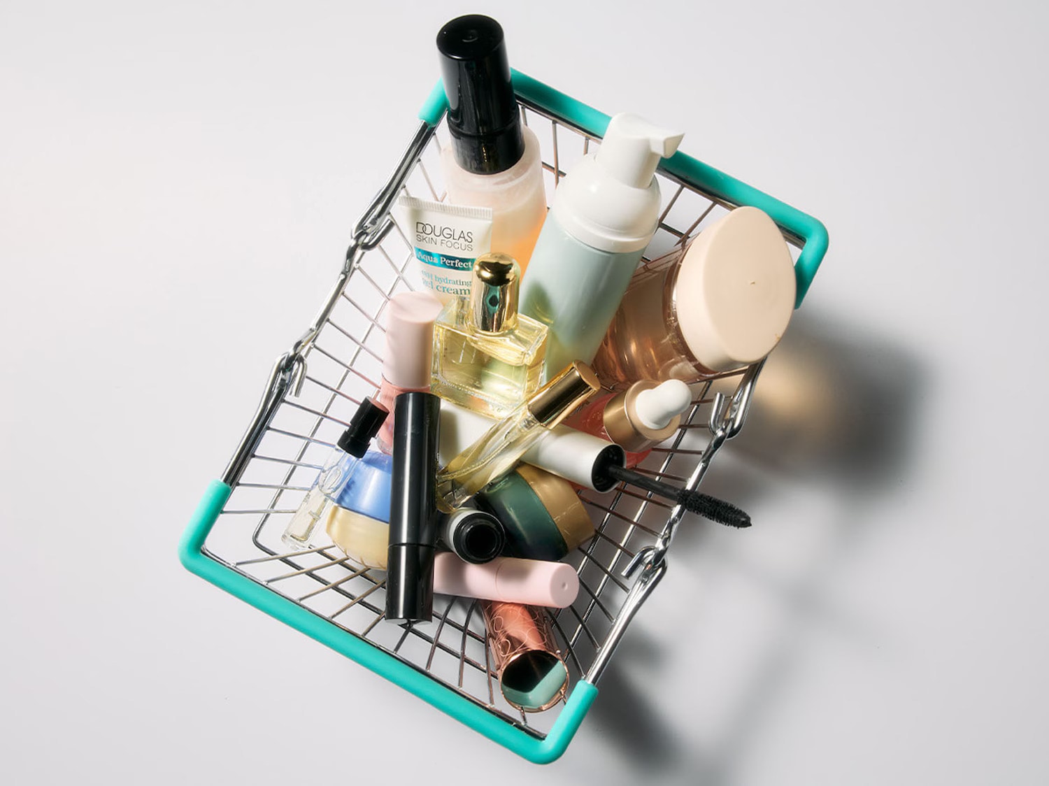 Pirkinių krepšelyje sudėti įvairūs kosmetikos produktai, įskaitant „Douglas Skin Focus Aqua Perfect“ drėkinamąjį kremą, kvepalus, tušą ir kitas grožio priemones.