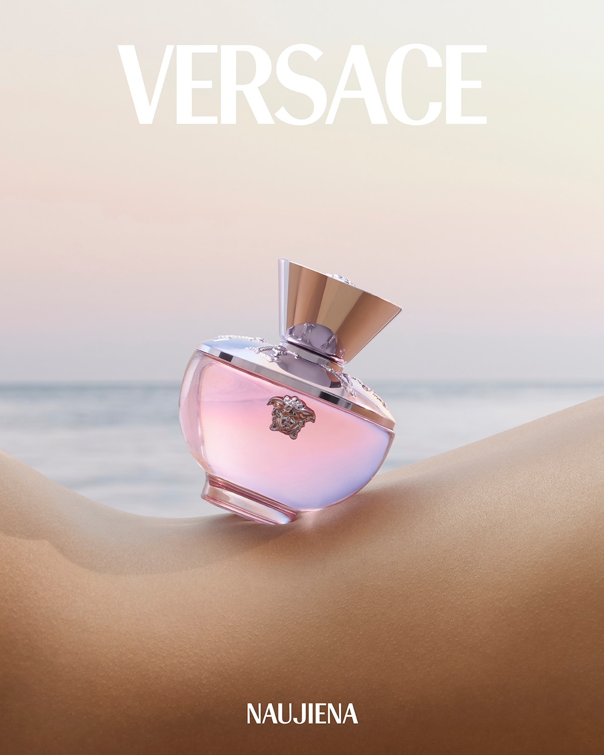 Versace aromatas su rožiniu skysčiu stikliniame buteliuke su aukso spalvos dangteliu, padėtas ant odos fono, viršuje užrašas „VERSACE“, apačioje – „NAUJIENA“.