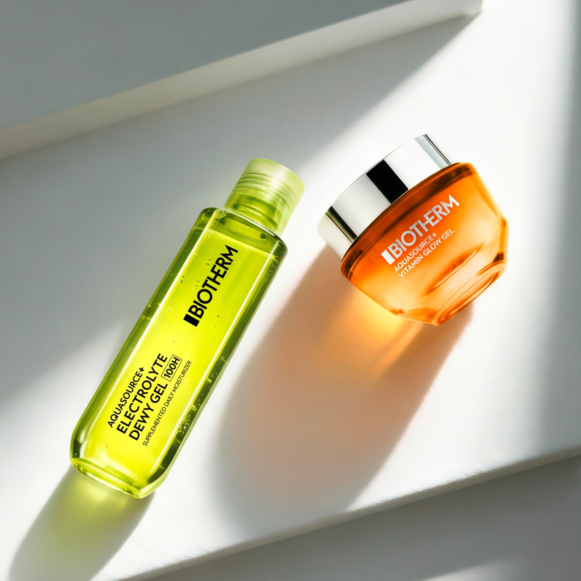 „Biotherm Aquasource+ Electrolyte Dewy Gel 100H“ papildytas kasdienis drėkiklis ir „Biotherm Aquasource+ Vitamin Glow Gel“ yra šalia, ant balto paviršiaus.