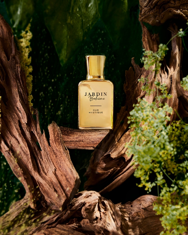 Auksinis „Jardin Bohème Oud Mystique“ kvepalų buteliukas, apsuptas žalios lapijos ir medienos, sukuria prabangų ir natūralų įvaizdį.