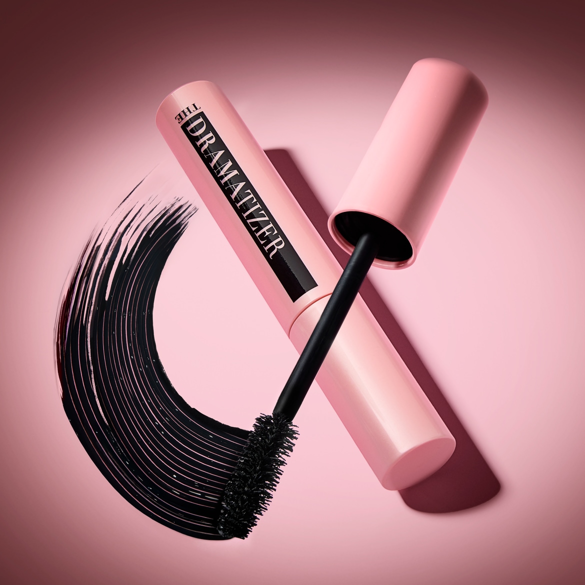 Rožinės spalvos „The Dramatizer“ tušo tūbelė su juodu šepetėliu ir juodu tušo brūkšniu ant rausvo fono.
