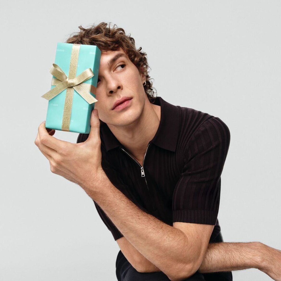 Vyras su garbanotais plaukais laiko dovaną, supakuotą į turkio spalvos popierių su auksiniu kaspinu, uždengdamas ja dalį veido.