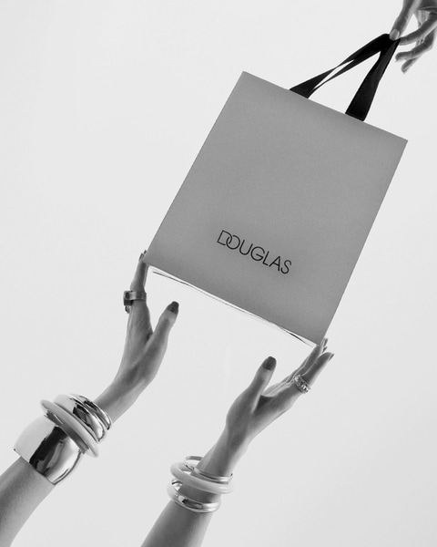 Sidabro spalvos „Douglas“ pirkinių krepšys su juodomis rankenomis, kurį laiko dvi rankos su sidabrinėmis apyrankėmis.