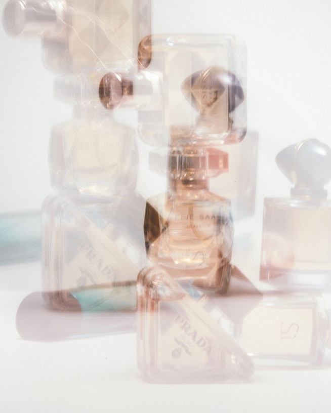 Švelniai atspalvinti kvepalų buteliukai, įskaitant „Prada“ ir „Armani“, išdėstyti ant balto paviršiaus, sukuria svajingą, eterinę kompoziciją.'