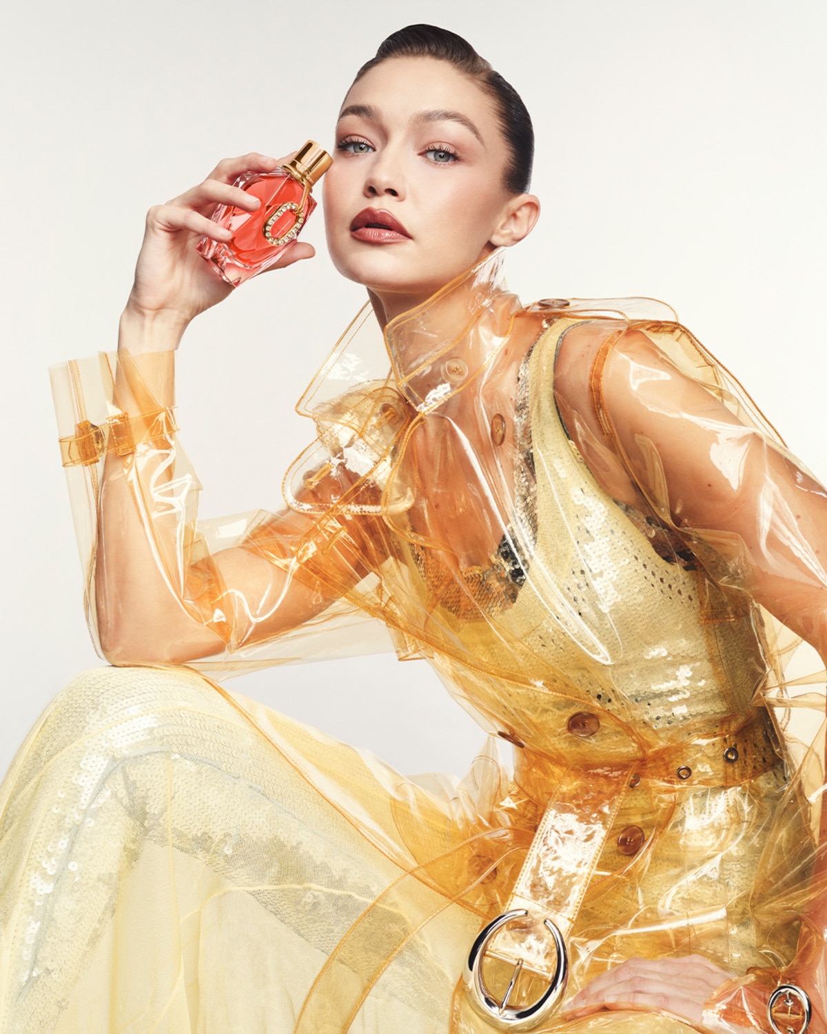 Šviesiaplaukė moteris su skaidriu oranžiniu lietpalčiu laiko rausvą kvepalų buteliuką su aukso spalvos dangteliu.