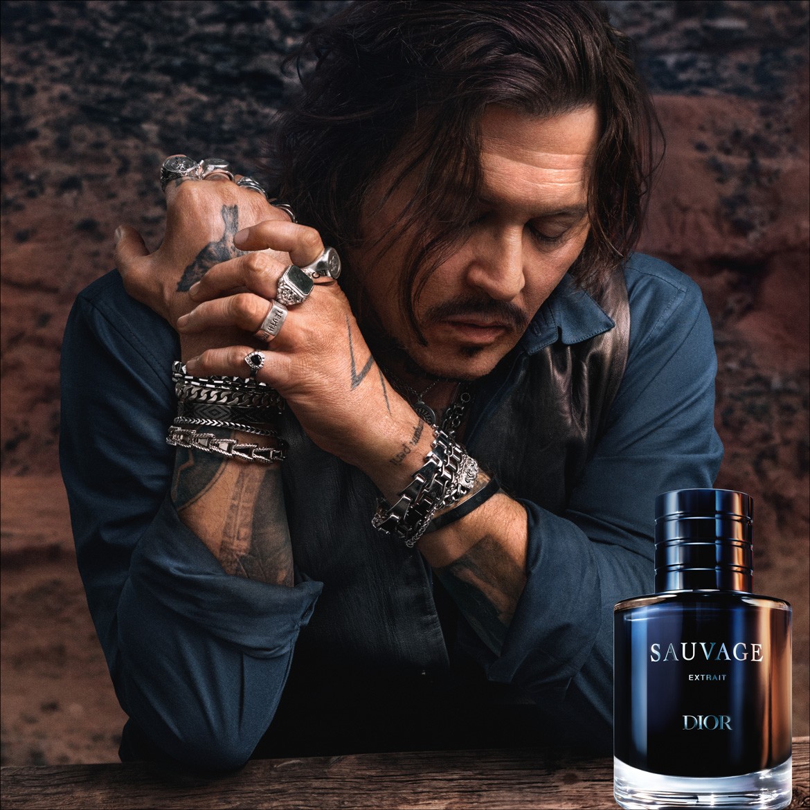 Johnny Deppas, pasipuošęs tamsiai mėlynais marškiniais ir odine liemene, su daugybe sidabrinių žiedų ir apyrankių, švelniai nuleidęs galvą, o šalia jo – tamsiai mėlynas „Dior Sauvage Extrait“ buteliukas su sidabriniu užrašu.