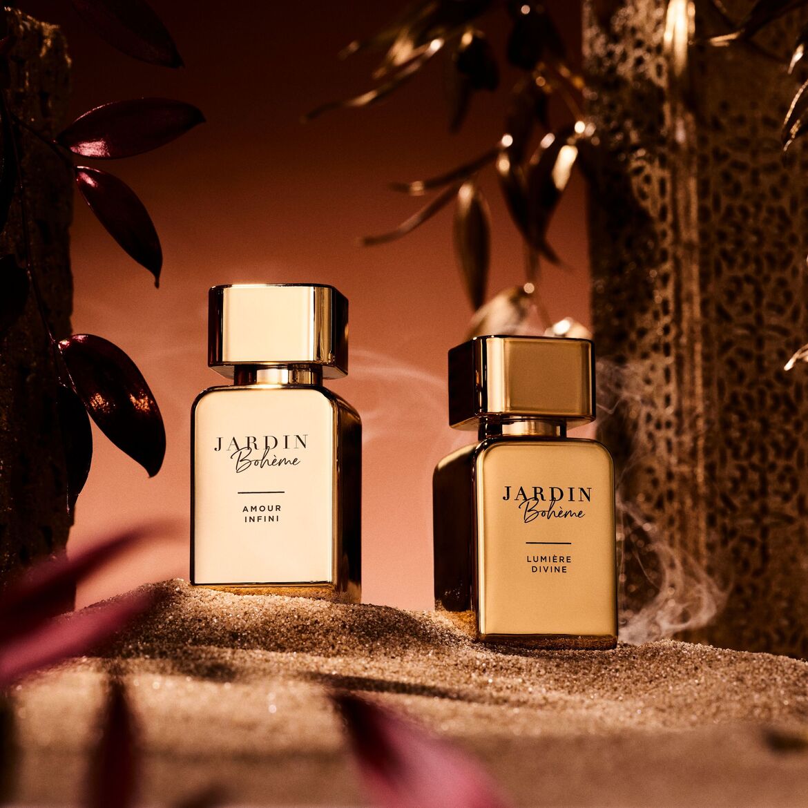 Du auksiniai „Jardin Bohème“ kvepalų buteliukai, vienas pažymėtas „Amour Infini“, kitas – „Lumière Divine“, elegantiškai eksponuojami ant smėlio, apsupti auksinių lapų.