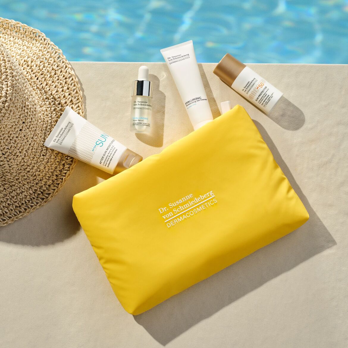 Šalia baseino guli geltonas kosmetikos krepšys su „Dr. Susanne von Schmiedeberg DERMACOSMETICS“ užrašu, apsaugos nuo saulės kremas SPF 50, hialurono gelinis valiklis, serumas ir losjonas po saulės.