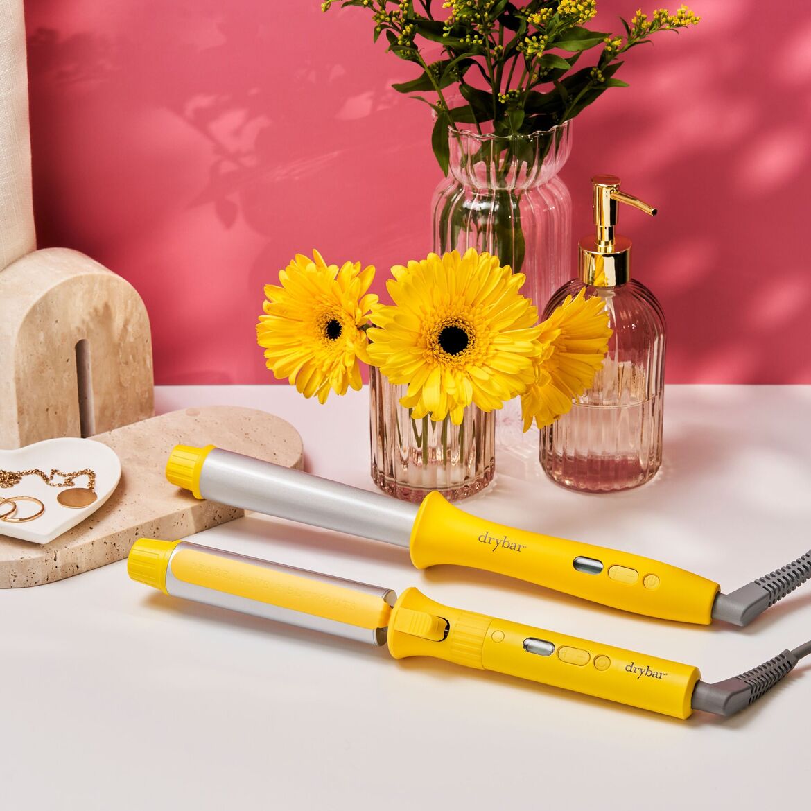 Du ryškiai geltoni „Drybar“ plaukų garbanojimo įrankiai su sidabriniais cilindrais ir juodais mygtukais guli ant balto paviršiaus, o fone – rožinė siena, gėlės ir dekoratyviniai elementai.