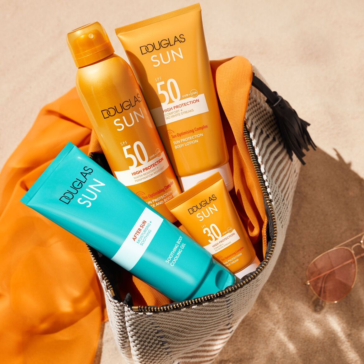 DOUGLAS SUN saulės priežiūros produktai (SPF 50 losjonas, purškiamas apsauginis kremas, SPF 30 veido kremas ir po saulės vėsinantis gelis) sudėti atviroje kosmetinėje ant smėlio šalia akinių nuo saulės.