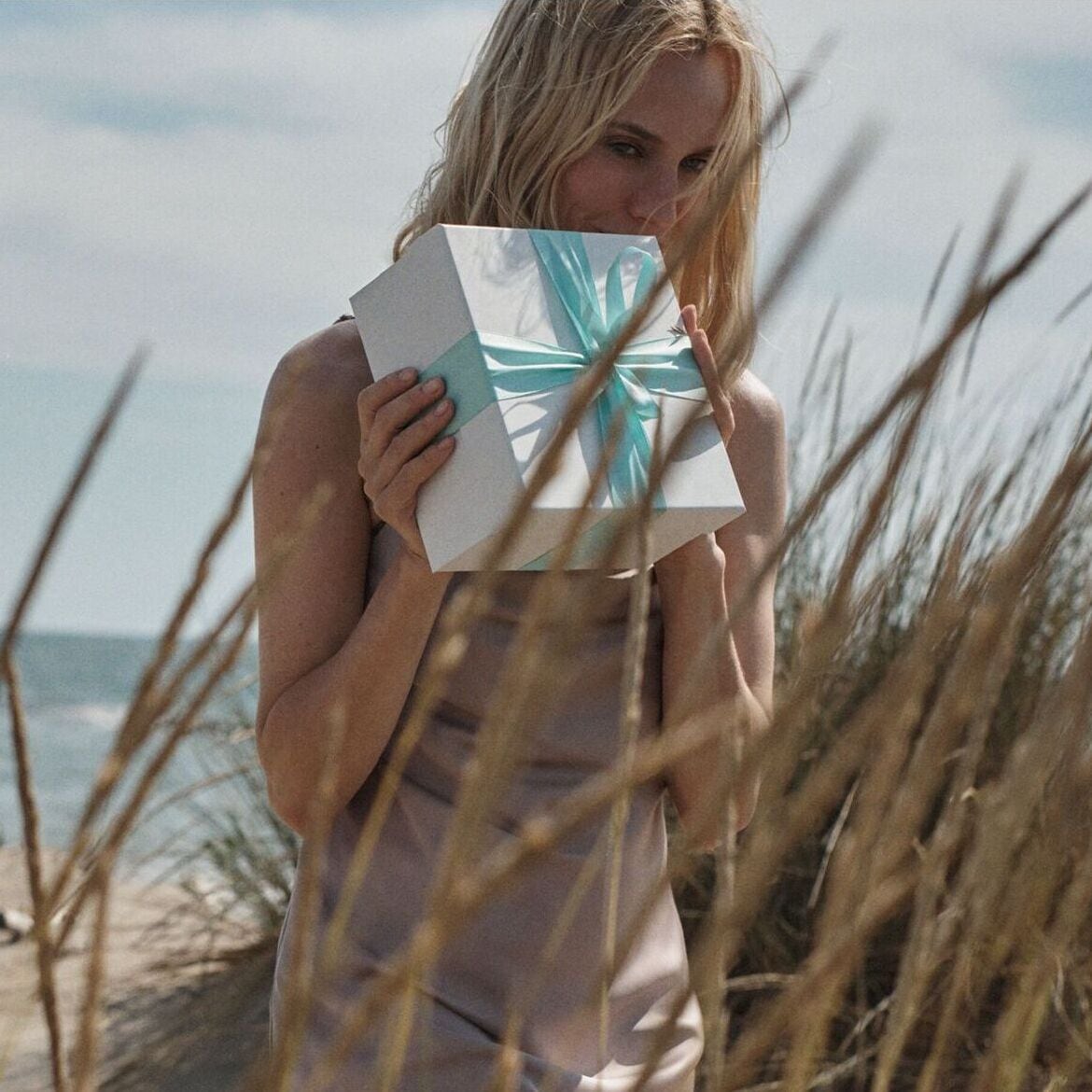 Šviesi blondinė laiko baltą dovanų dėžutę, perrištą žydru kaspinu, smėlėtoje paplūdimio aplinkoje.