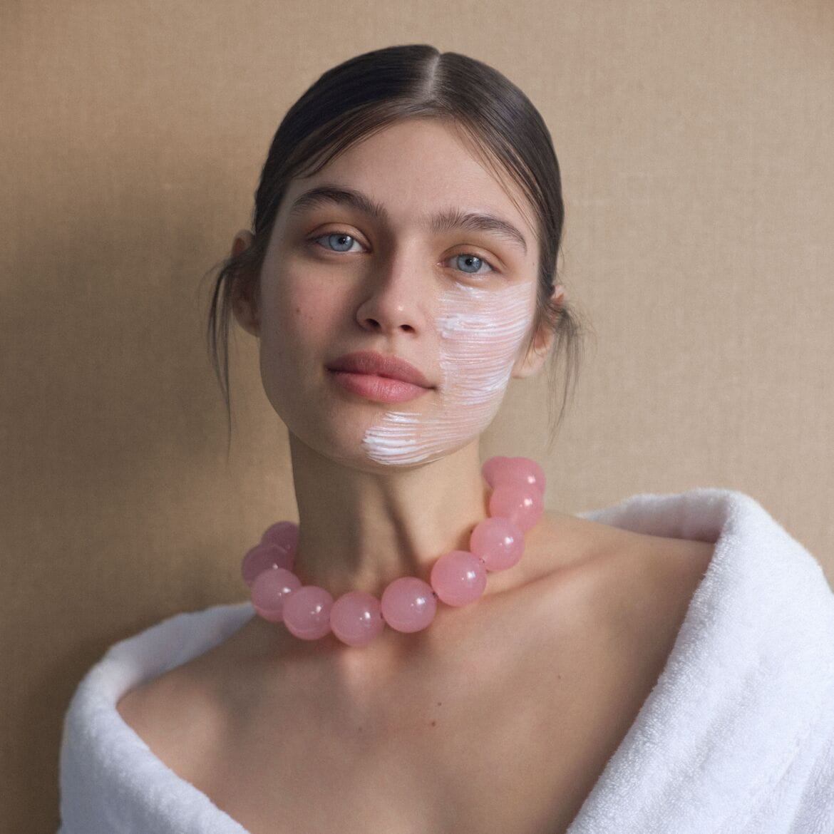 Jauna moteris su balta veido kauke ant skruosto, dėvinti chalatą ir rožinių karoliukų vėrinį, pasiruošusi odos priežiūros ritualui.