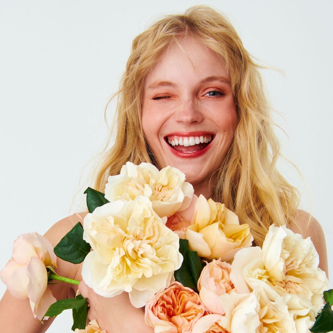 Šviesiaplaukė moteris su šypsena ir gėlėmis rankose, vienu akies mirktelėjimu, spinduliuoja džiaugsmą ir gaivumą.