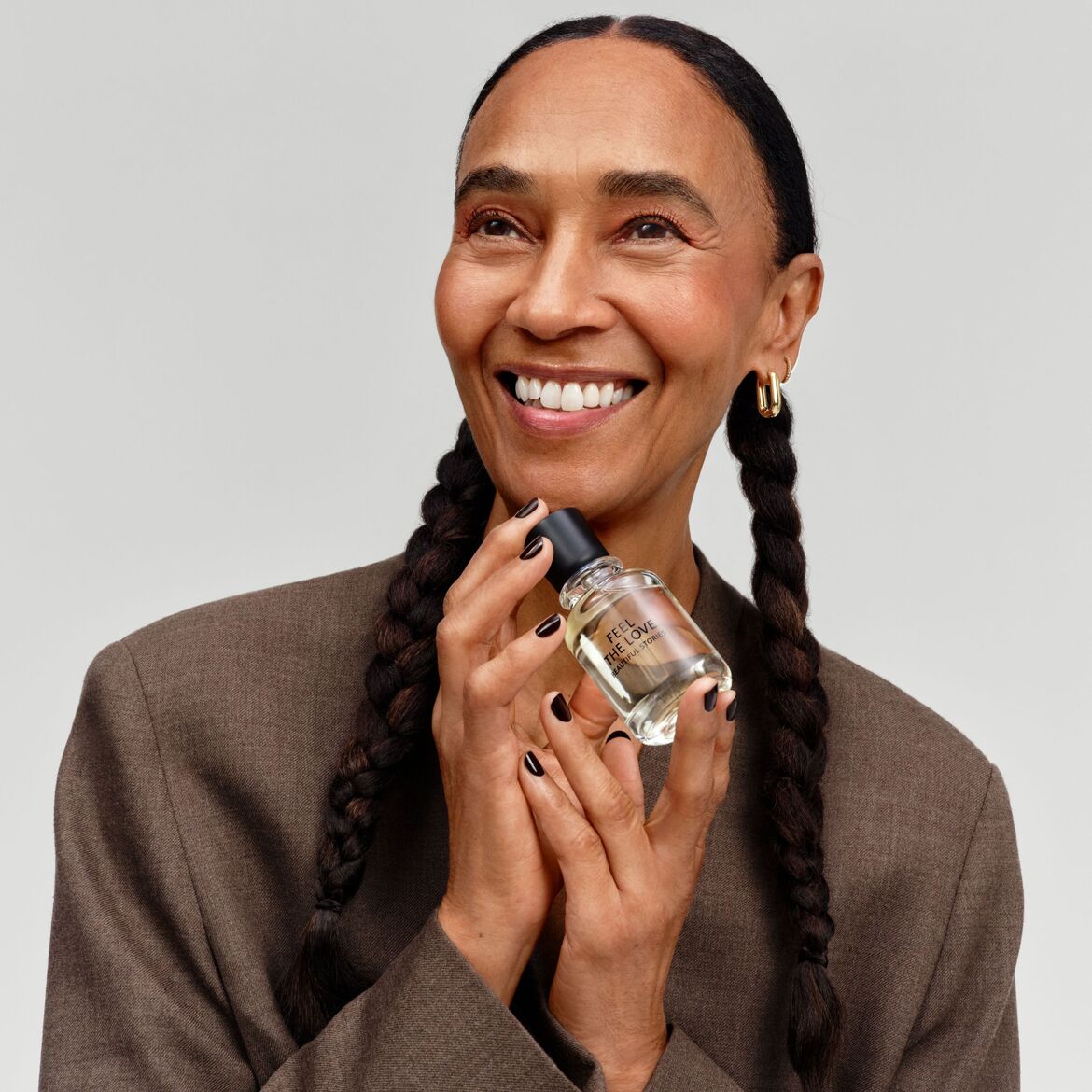 Švytinti moteris su rudu švarku laiko skaidrų kvepalų buteliuką su juodu dangteliu, ant kurio užrašyta „Pajusk meilę“.