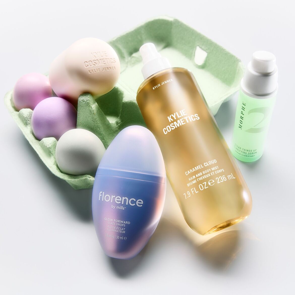Įvairūs kosmetikos gaminiai, įskaitant „Kylie Cosmetics“ ir „Florence by Mills“, išdėstyti ant žalio kiaušinių dėklo su spalvotais kiaušiniais, sukuria šventinę ir spalvingą kompoziciją.