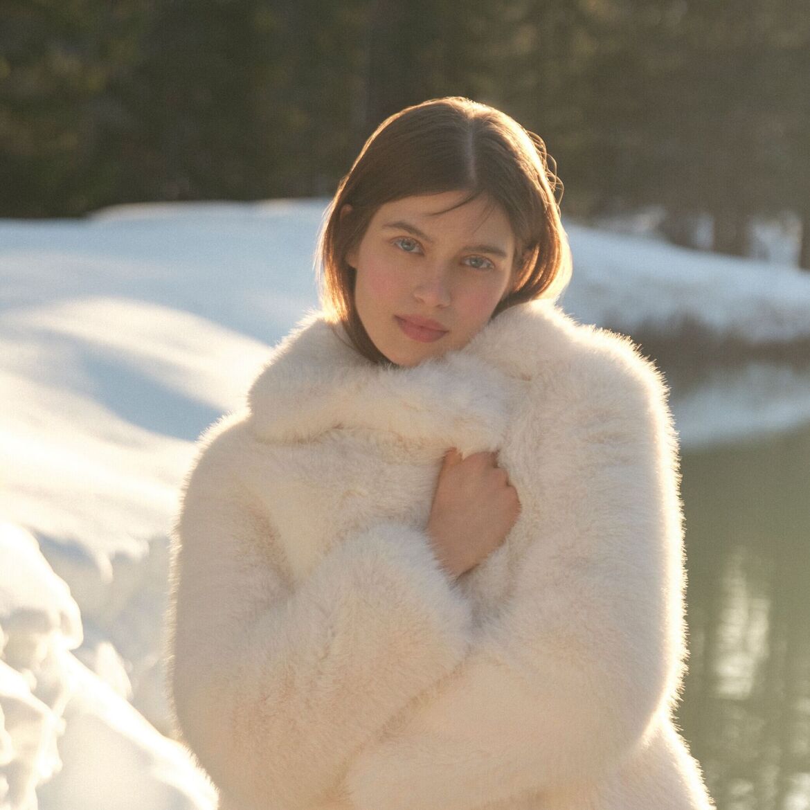 Moteris su baltu kailiniu paltu snieguotame kraštovaizdyje, spinduliuojanti šilumą ir stilių.
