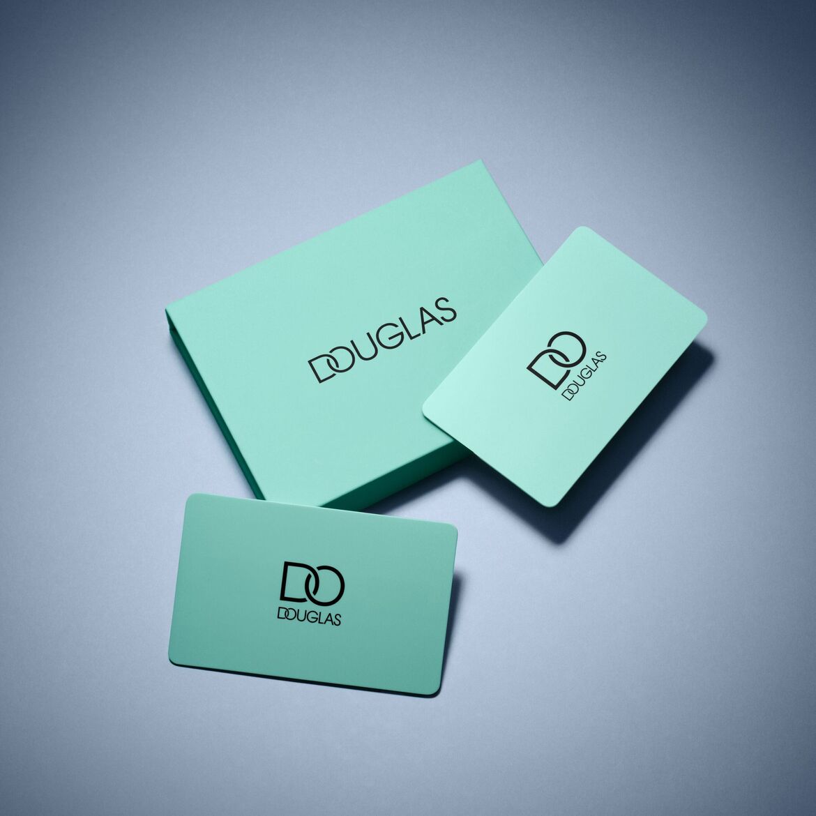 Šviesiai mėlynos spalvos „Douglas“ dovanų dėžutė ir dvi dovanų kortelės su prekės ženklo logotipu ant pilko paviršiaus.