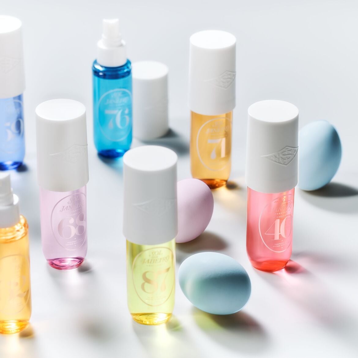 Įvairių spalvų „Sol de Janeiro“ purškikliai su baltais dangteliais ir pasteliniais kiaušiniais, išdėstyti ant balto paviršiaus.